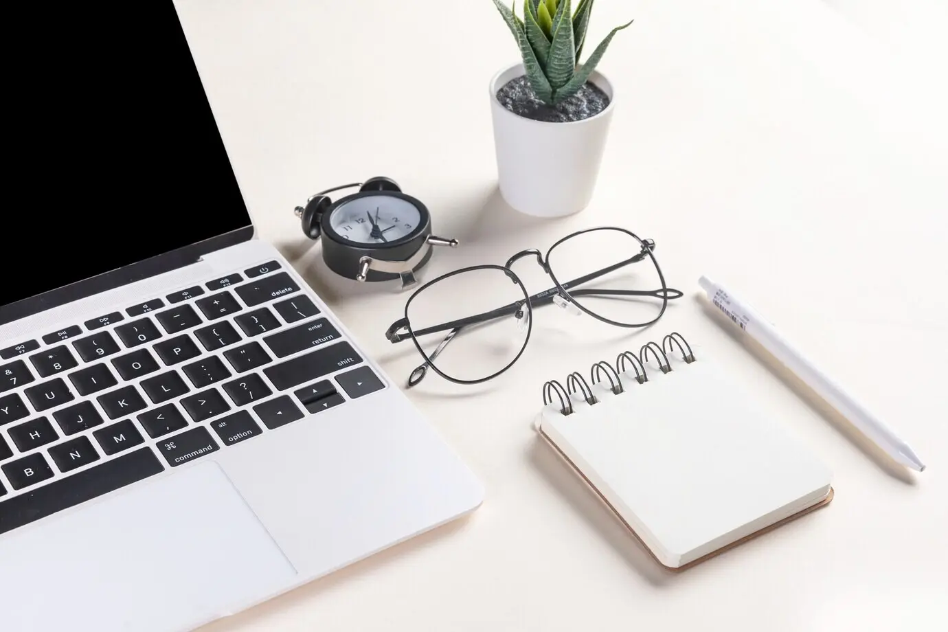 A close-up view of an open laptop, glasses, a plant, a pen, a notepad, and an alarm clock.