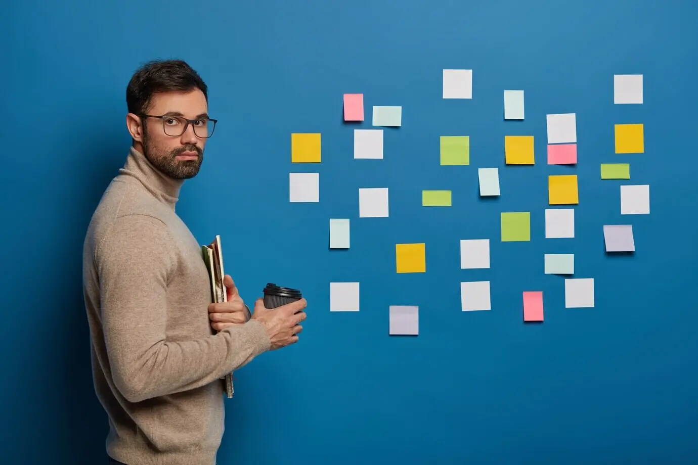 Bearded male student takes a coffee break indoors, holds a disposable cup with a hot beverage, and uses stickers for remembering new information.
